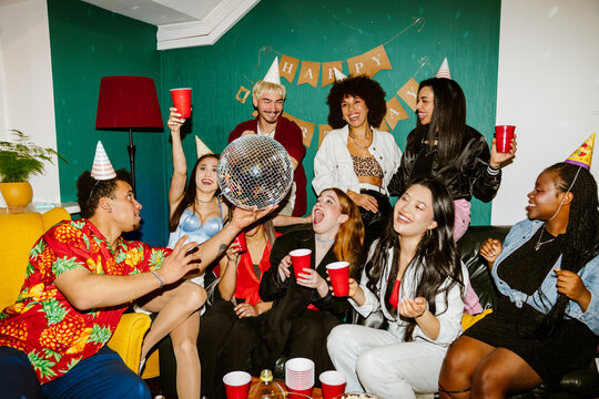 A vibrant multinational group of young people celebrating a birthday party, wearing party hats, enjoying drinks, playing with a disco ball, in a festive room decorated with colorful items and lights.
