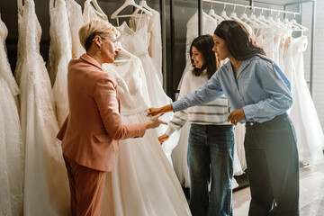 A Black bride in a blue blouse, her White female friend, a White female consultant with short hair, shopping for wedding dresses together in a bridal store, surrounded by elegant white gowns.