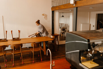 A White female cafe owner rearranging chairs and preparing a table during a renovation of her cozy cafe, focusing on the interior decor with a modern design and clean wooden furniture.