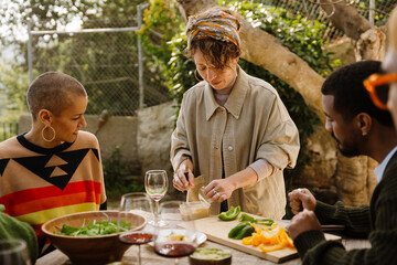 A multinational group of adult people are preparing and enjoying dinner together outdoors, chopping vegetables and sharing a moment with wine and surrounded by greenery in a warm, relaxed atmosphere.