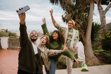 Young multiracial friends, including two women and two men, smiling and taking a selfie while posing playfully outdoors on a terrace with scenic views in the background.