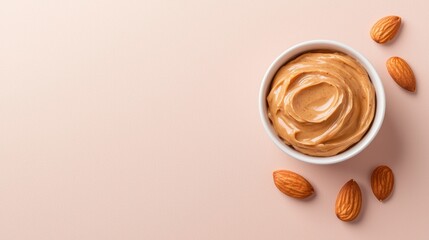 Flat lay of a small white bowl filled with a creamy peanut butter. the bowl is placed on a light pink background and there are several whole almonds scattered around it.