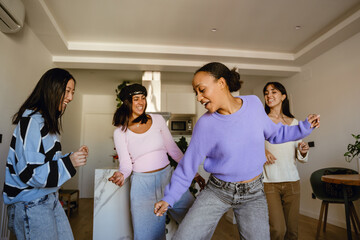 A group of diverse young women, including Black, Asian, and Hispanic friends, dancing and smiling in a modern apartment living room during the daytime.