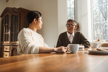 An Asian adult man talking with his elderly grandfather at a wooden table in a bright home interior, sitting with coffee mugs in a warm and relaxed atmosphere.
