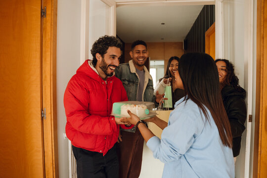 A group of diverse young friends arrive at an apartment for a birthday celebration. One man hands a cake to a woman at the door as others smile and carry gifts indoors. - Powered by Adobe