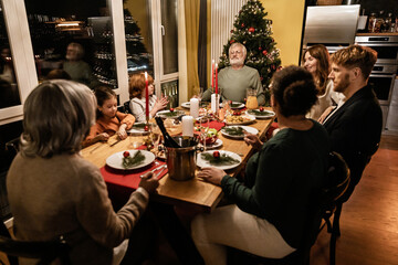 A multiracial family of adults and children sit around a decorated dinner table at night and share a festive Christmas meal near a lit tree in a cozy indoor kitchen.