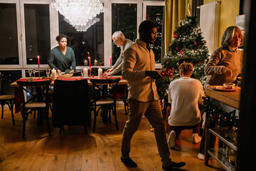 Obraz premium A multinational group of people of different ages enjoying a Christmas dinner, preparing food, with the Christmas tree, and sharing festive moments together in a cozy home with candles and decor.