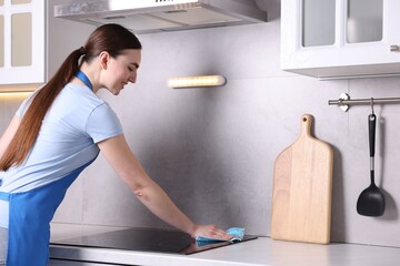 Smiling woman cleaning induction cooktop with rag in kitchen