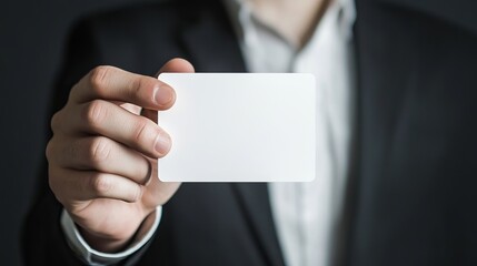 Fototapeta premium A person in a suit holding a blank white card in their hand against a dark background in a studio