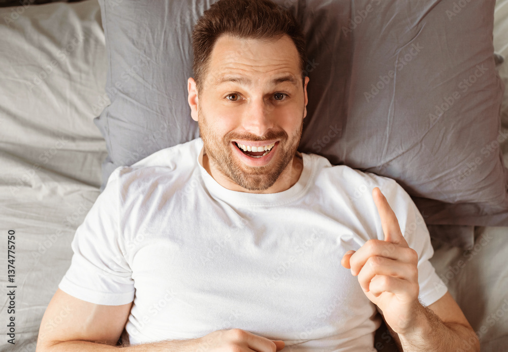 Poster excited man having idea after good sleep pointing finger up looking at camera lying in bed in bedroo