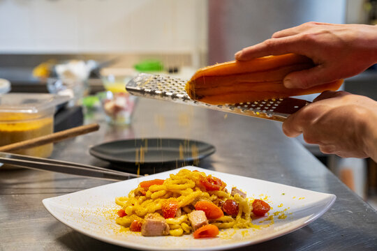 Chef grating bottarga over a plate of fresh pasta with cherry tomatoes in a professional kitchen. Gourmet Italian seafood dish being finished with traditional Mediterranean ingredients