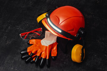 An orange hard hat with a headlamp and ear protection is placed alongside orange gloves and safety goggles on a dark textured surface. © vadish