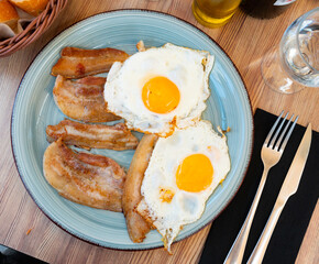 Plate is filled with cooked homemade food. Fried eggs cooked with slices of fatty pork bacon. Sliced bread, plate of food, cutlery and glasses are placed on wooden table
