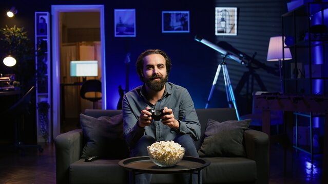 Excited man holding controller in neon illuminated apartment, playing videogames. Happy gamer on couch using joystick to participate in PvP game on console system, relaxing and having fun, camera B