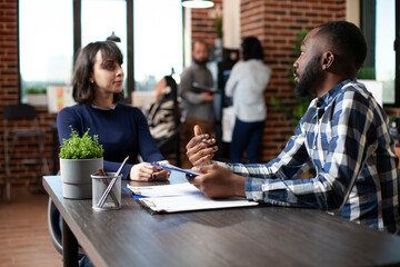 Young female job candidate listening attentively to african american office worker, discussing recruitment process at startup office. HR male representative with clipboard, interviewing a white woman.