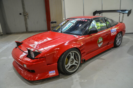Heavily modified red Nissan 200SX S13 with large rear wing and aggressive body kit. The car features aftermarket wheels lowered suspension, built for performance and show