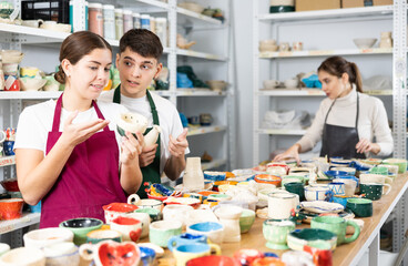 Fototapeta premium Interested young man and women in aprons viewing handmade clay products on the table in pottery workshop