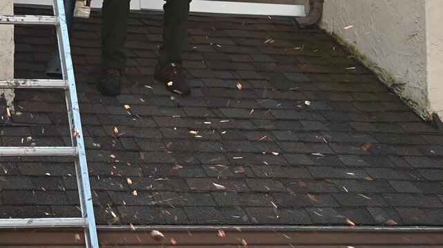 Senior man on asphalt shingle roof cleaning off pine needles and tree debris with leaf blower, property maintenance
