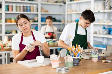 Interested young man and woman in pinafores sculpting handmade jars of potter-earth material in art...