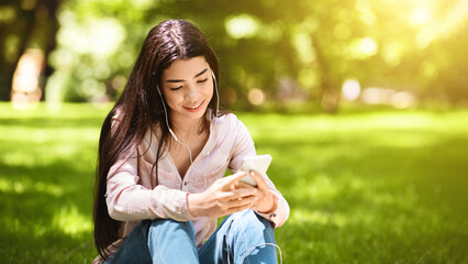Dreamy Asian Girl Listening Her Favorite Songs With Smartphone And Earphones Outdoors, Sitting On Lawn In Park, Enjoying Music