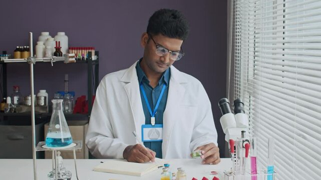 Serious Indian lap worker sitting at work desk taking notes while looking at tiny bottle of vaccine