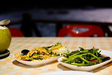 Thai dishes of stir-fried morning glory (Kang Kong) and red curry chicken served with a fresh young coconut at Cliff Restaurant, Patok Bay, with a night view of the sea in the background - Koh Racha