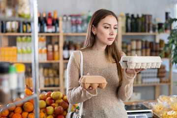 Thoughtful smiling young woman in cozy grocery store holding two different egg cartons, comparing options before deciding to buy