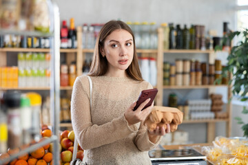 Thoughtful young woman making purchases in farmer grocery store, using smartphone to scan barcode on carton box with fresh chicken eggs. Modern shopping concept