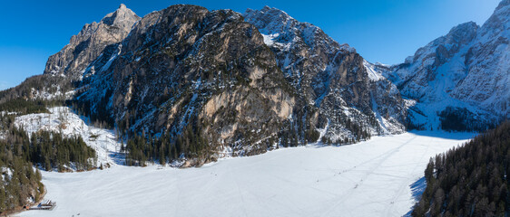 Frozen Braies Lake in northern Italy, surrounded by snow dusted mountains and forests. Tracks and figures cross the snowy surface near a wooden dock.