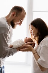 Portrait of a young family with infant in his arms photographed on a light window background.
