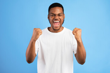Fototapeta premium Winner. Emotional Black Guy Shouting Yes Shaking Fists Looking At Camera Standing On Blue Background. Studio Shot