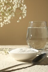 Collagen powder in bowl, spoon, glass of water and gypsophila branches on light textured table against beige background, closeup