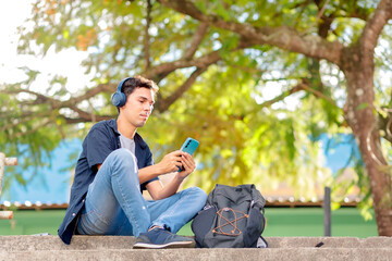 Obraz premium Young Brazilian man sitting outdoors on university steps, wearing wireless headphones and using a smartphone. Concept of digital lifestyle, online communication, and college student life.