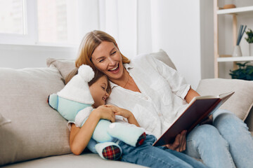 Mother and daughter reading a book together on a cozy couch while cuddling a teddy bear in a warm home setting
