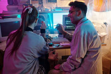 Two diverse young programmers chatting and smoking while sitting at wooden desk with vintage computer monitors and tape recorder on it