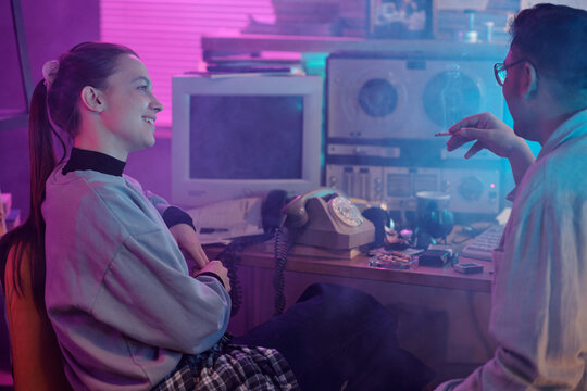 Programmer holding cigarette talking with smiling girl while they sitting in front of wooden desk cluttered with tools and equipment