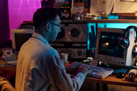 Modern male Indian hacker decoding data at wooden table with retro computer displaying running code, copy space