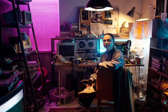 Caucasian young woman posing for camera while sitting on chair in dimly lit messy room with vintage tech equipment