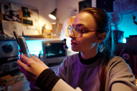 Portrait of Caucasian young woman using old cellphone while sitting on chair in dimly lit messy room with vintage tech equipment