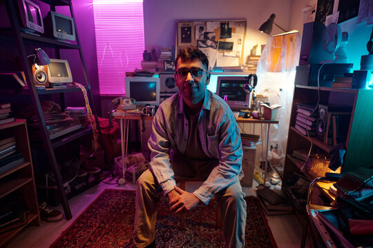 Young Indian man sitting in cluttered room, filled with retro computers and devices, with neon lighting
