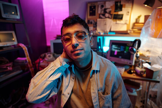 Portrait of young male programmer wearing glasses posing for camera in dimly lit room with retro devices and notice board in background