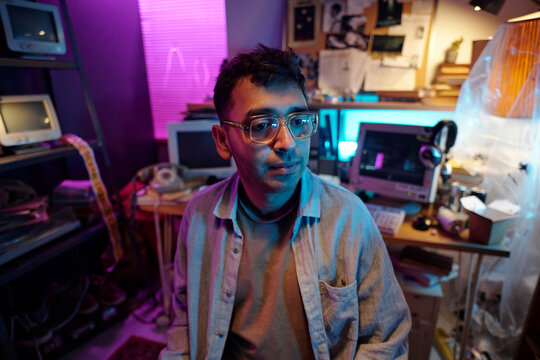 Medium closeup of young programmer wearing glasses sitting in dimly lit room with retro devices and notice board in background