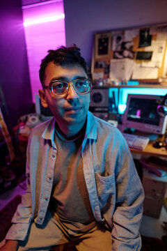 Vertical medium closeup of young hacker wearing glasses posing for camera in dimly lit room with retro devices in background