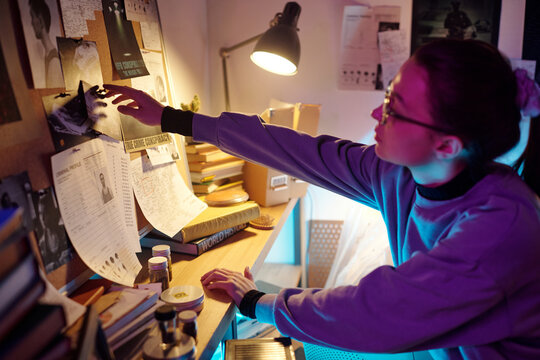 Young curious amateur investigator getting up from her chair to remove picture from pin board, then sitting down and examining it