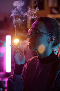 Vertical close-up of young woman with glasses and hair in ponytail smoking cigarette in dimly lit room