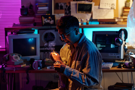 Young Indian man with glasses and hair in ponytail lighting up cigarette in dimly lit room using match