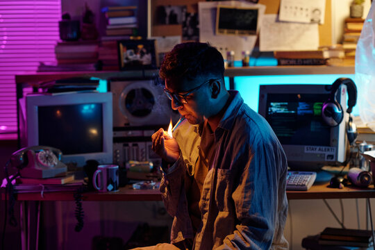 Modern Indian man with glasses lighting up cigarette in dimly lit cluttered room using match