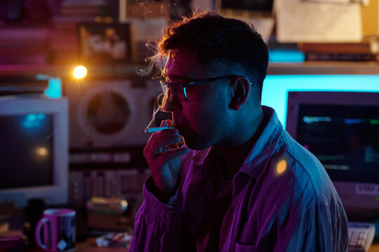 Close-up view of young male hacker with glasses taking drag from cigarette while working in dimly lit room
