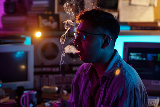 Young male hacker with glasses exhaling smoke through mouth while smoking in dimly lit room
