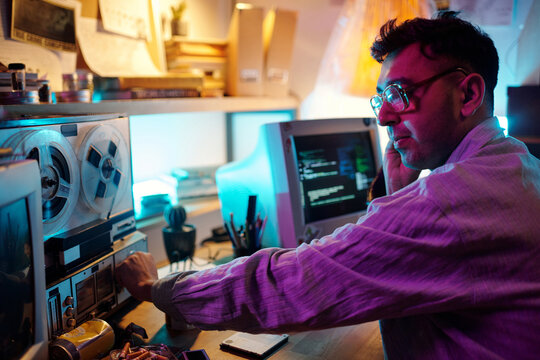 Young male hacker turning on retro tape recorder and listening to recording with headphones in room with neon light